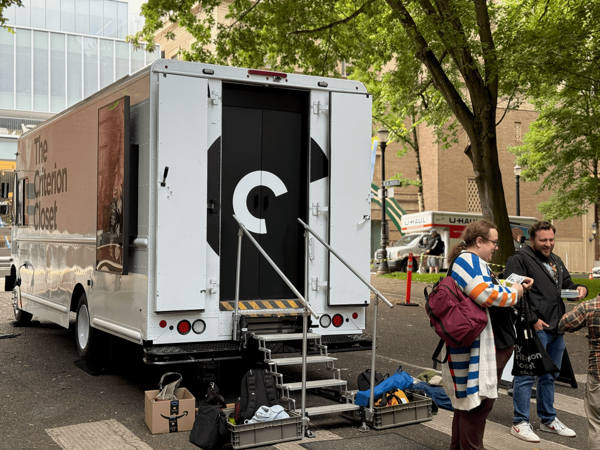 The back of the white Criterion Mobile Closet van. You can see a video screen on the left side, a couple of very happy people outside, and steps leading up to a mysterious black door with the Criterion C logo on it.